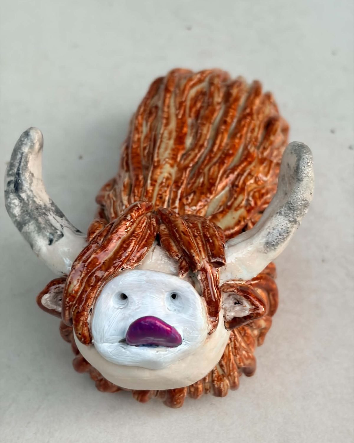 Murdo, a handmade ceramic Highland cow with a ginger coat, white face and a purple nose, mouth slightly open